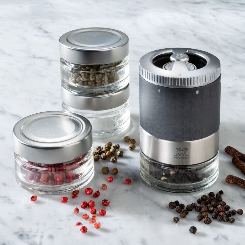 Drei Glasdosen für Pfeffer mit Edelstahldeckeln und eine kompakte Pfeffermühle, umgeben von bunten Pfefferkörnern.