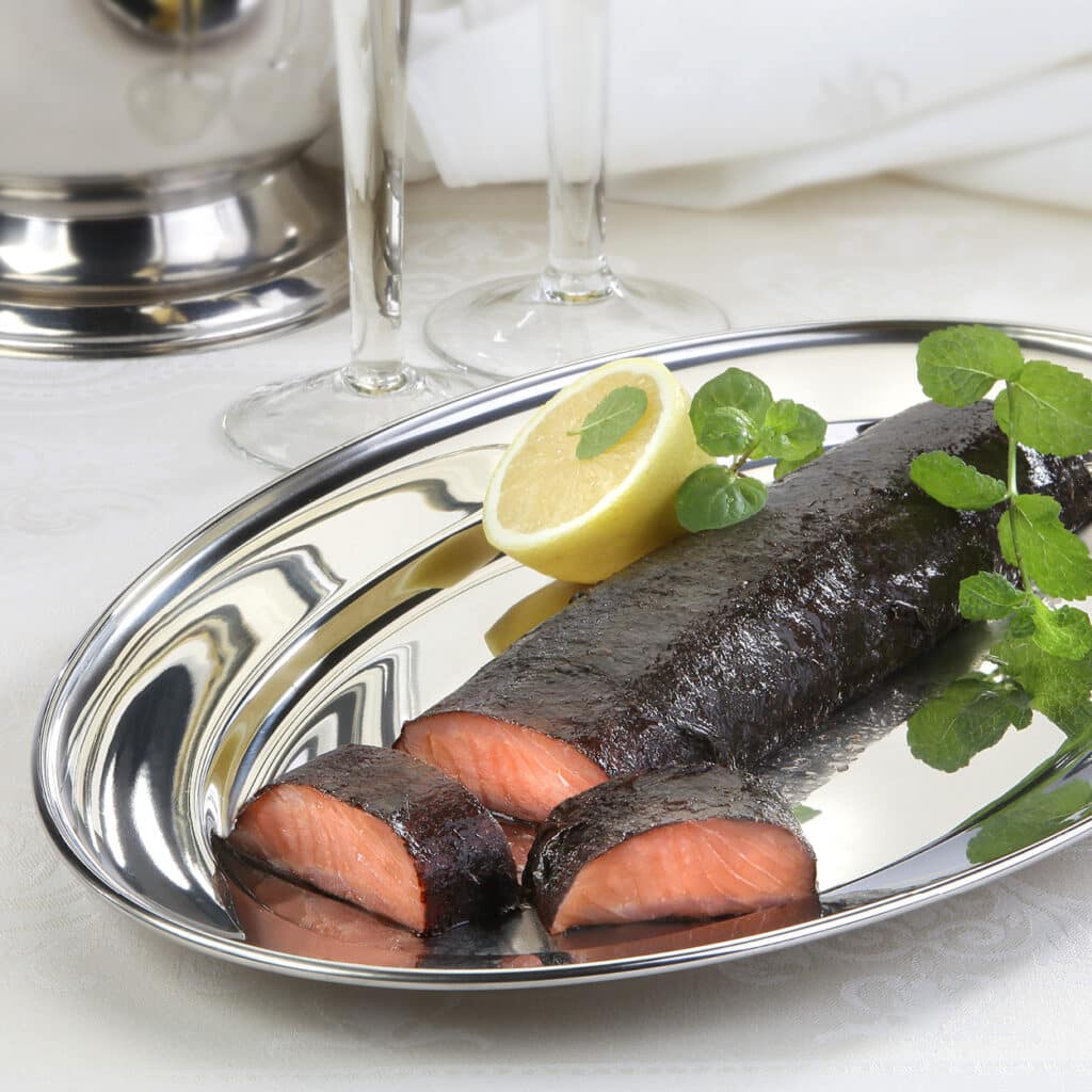Geräuchertes Lachsfilet im dunklen Kräutermantel, angerichtet auf einer silbernen Servierplatte mit Zitronenscheibe und frischen Kräutern.