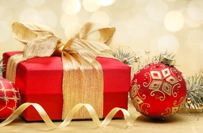 Rotes Weihnachtsgeschenk mit goldener Schleife und glitzernden Christbaumkugeln vor goldenem Bokeh-Hintergrund.