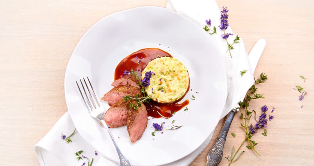 Magret de canard rosé tranché, servi avec une purée de légumes et une sauce brune, décoré d’herbes et de fleurs comestibles sur une grande assiette blanche.