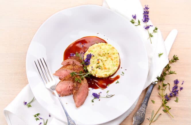 Magret de canard rosé tranché, servi avec une purée de légumes et une sauce brune, décoré d’herbes et de fleurs comestibles sur une grande assiette blanche.