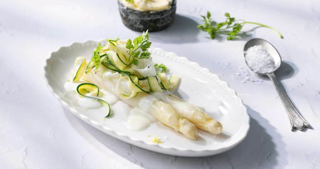 Asperges blanches servies avec des rubans de courgette, napp&eacute;es d&rsquo;une sauce cr&eacute;meuse et garnies d&rsquo;herbes fra&icirc;ches sur une assiette blanche.
