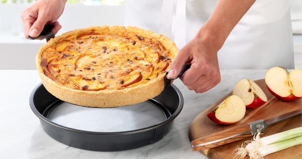 Une tarte sal&eacute;e aux pommes, dor&eacute;e et croustillante, est retir&eacute;e d&rsquo;un moule par deux mains. &Agrave; c&ocirc;t&eacute;, sur une planche en bois, se trouvent des pommes coup&eacute;es, un couteau et un poireau.