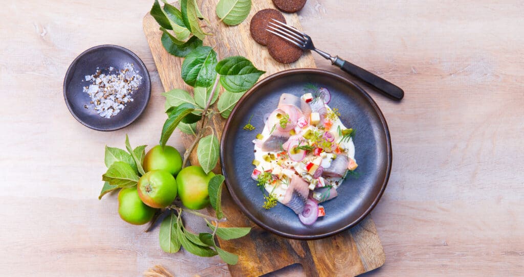 Un plat raffin&eacute; de poisson marin&eacute; servi dans un bol sombre, garni de l&eacute;gumes color&eacute;s et d&rsquo;herbes fra&icirc;ches. &Agrave; c&ocirc;t&eacute;, sur une planche en bois, reposent des pommes vertes avec leurs feuilles, du sel et une fourchette.